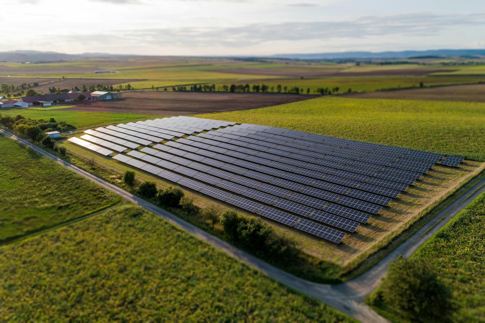 Vista aérea de un arreglo fotovoltaico entre parcelas de cultivo al final de la tarde, con las hileras de seguidores en diagonal y el sol bajo reflejándose sobre los módulos.